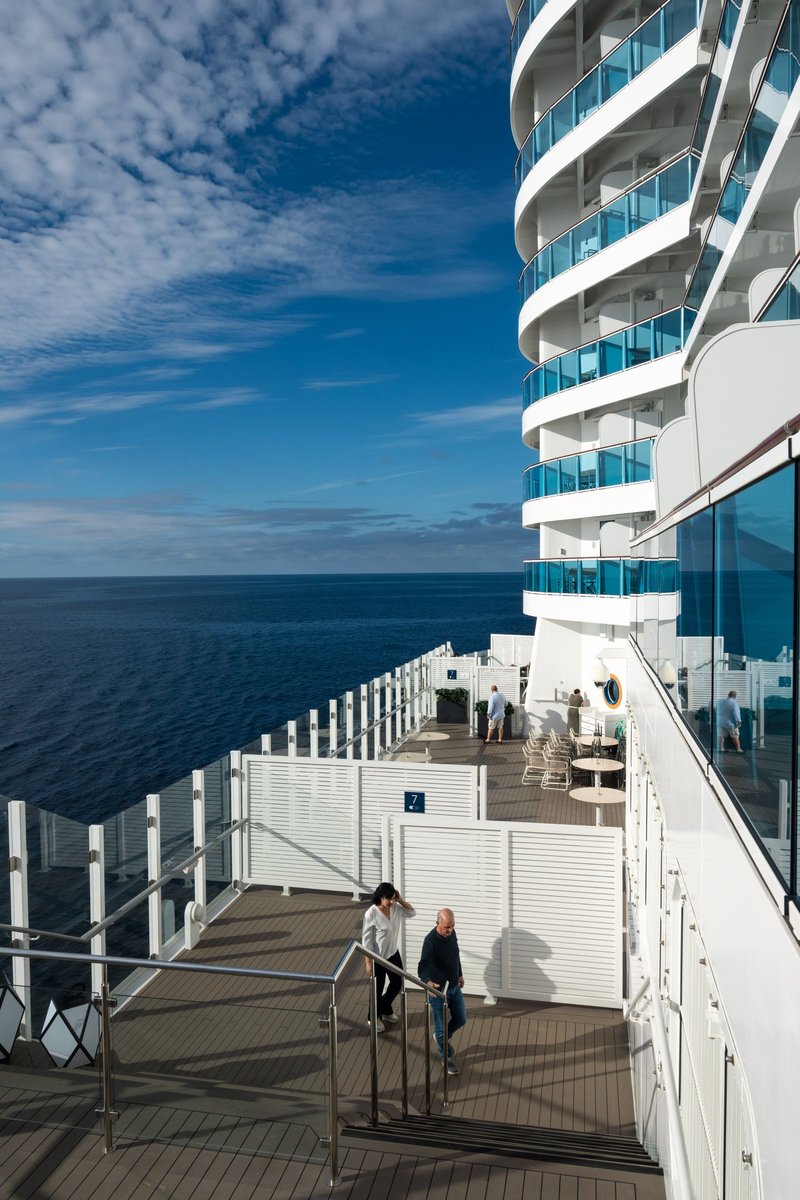 Passengers walking a sunny cruise-ship deck with glass balcony railings overlooking blue ocean