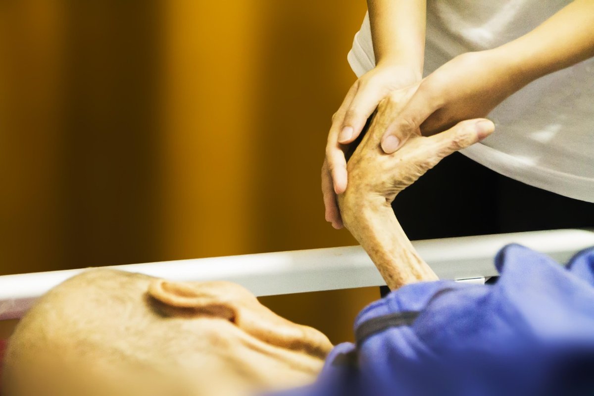A caregiver gently holding the hand of an elderly patient lying in bed